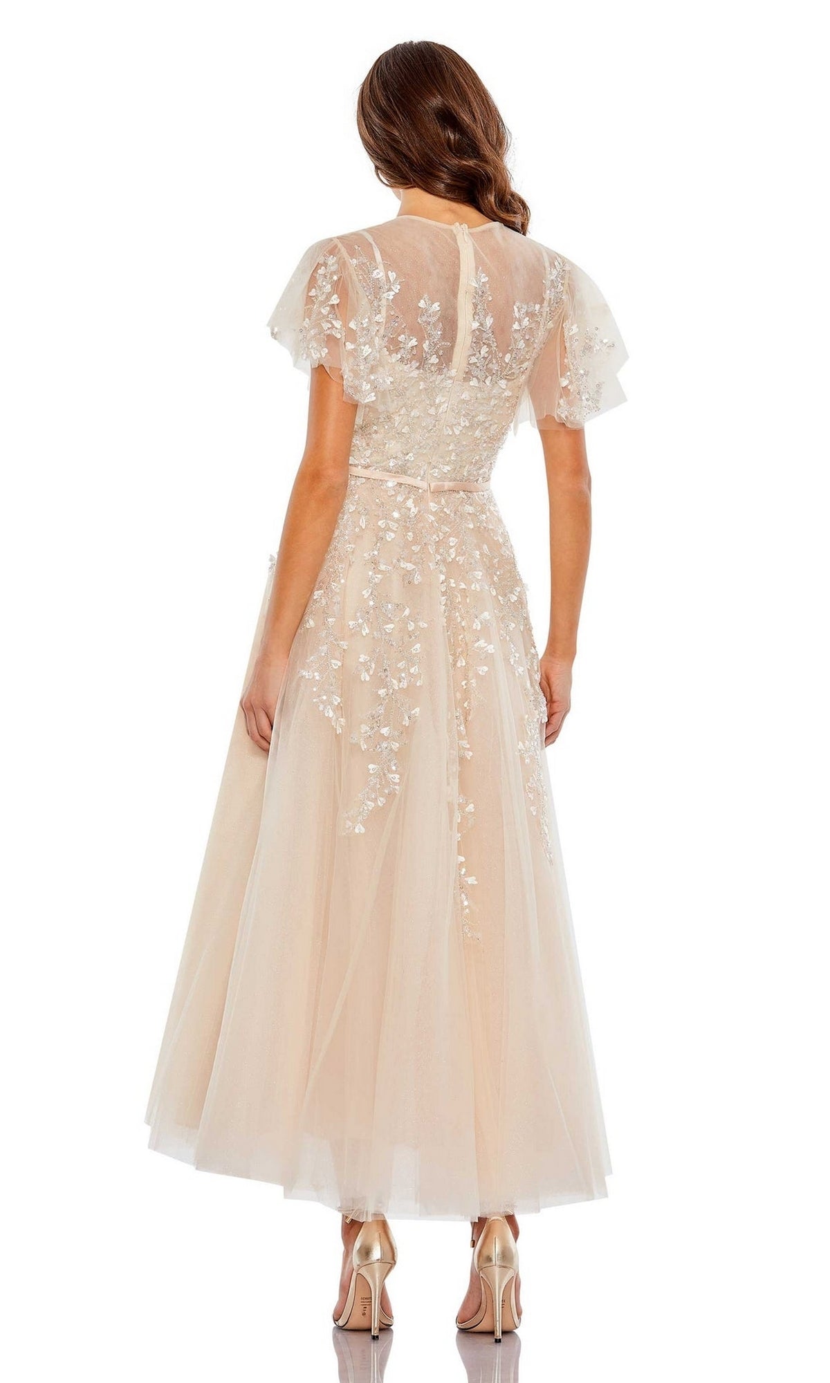 A woman with wavy hair stands in high heels, back to the camera, modeling the Mac Duggal Long Formal Dress 20362—an A-line, tea-length dress in light peach with floral embroidery and short sleeves.